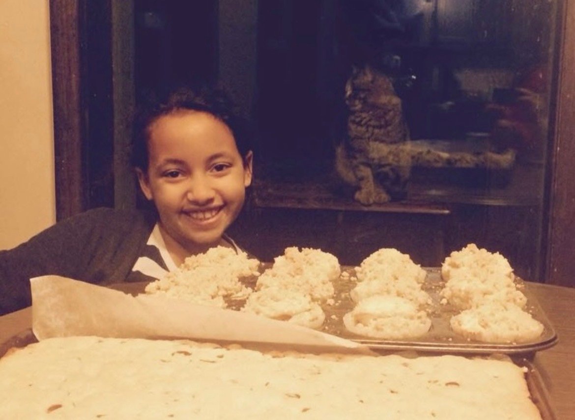 Evangeline smiling behind a tray of fresh streusel-topped muffins and a sheet cake